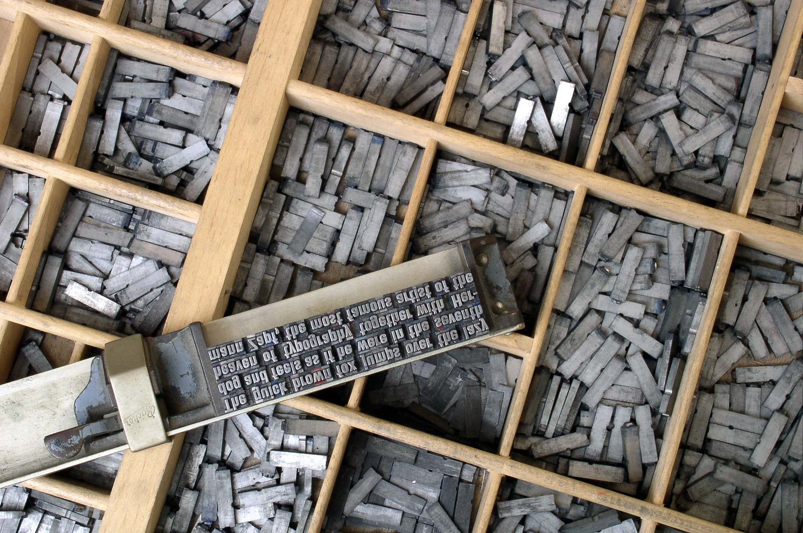 Separate metal sorts lie in wooden compartments beside a composing stick holding several lines of reversed letters.