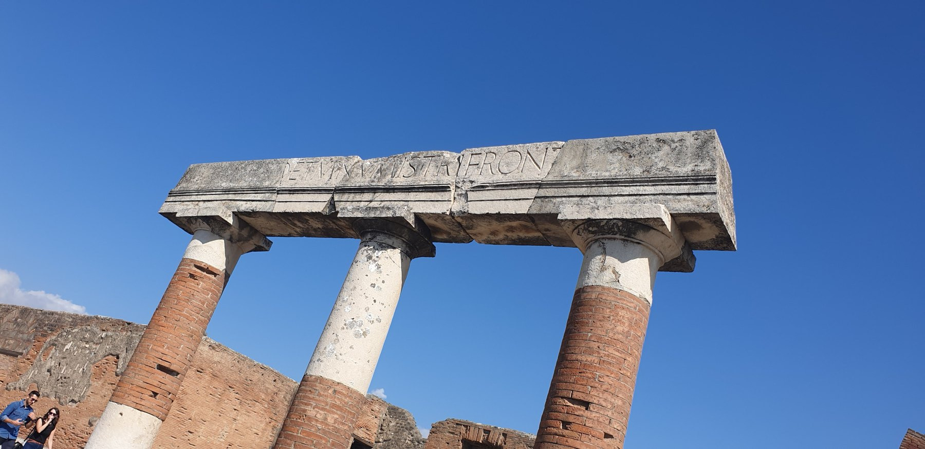 Three brick and stone columns support a fragmented stone entablature bearing part of a Latin inscription.