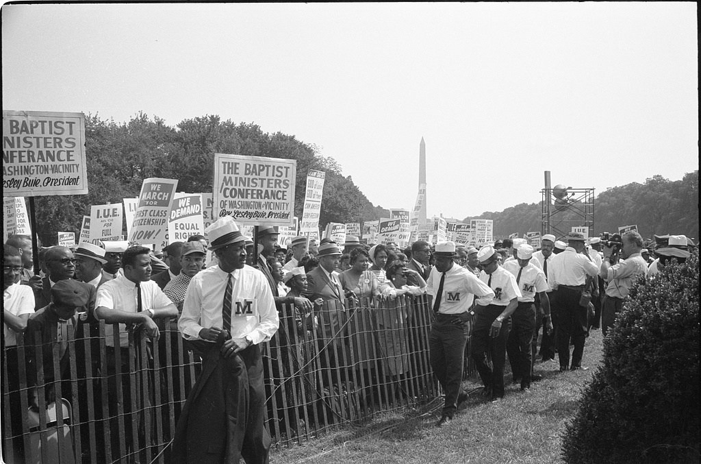 Marshals wearing M armbands stand beside a fence with marchers and civil rights placards behind them.