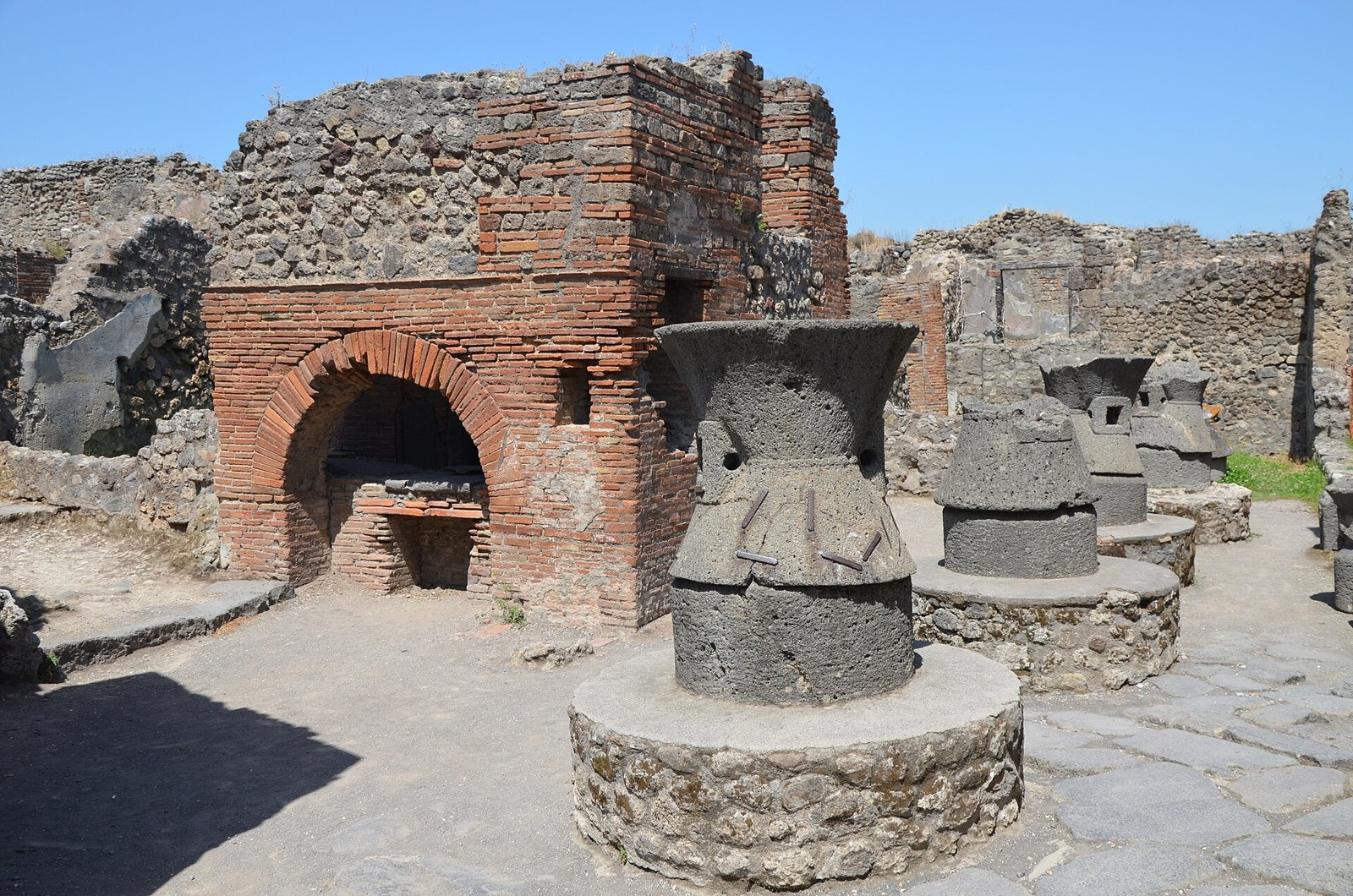 Large hourglass-shaped stone mill components stand beside the surviving masonry of a Pompeian bakery.