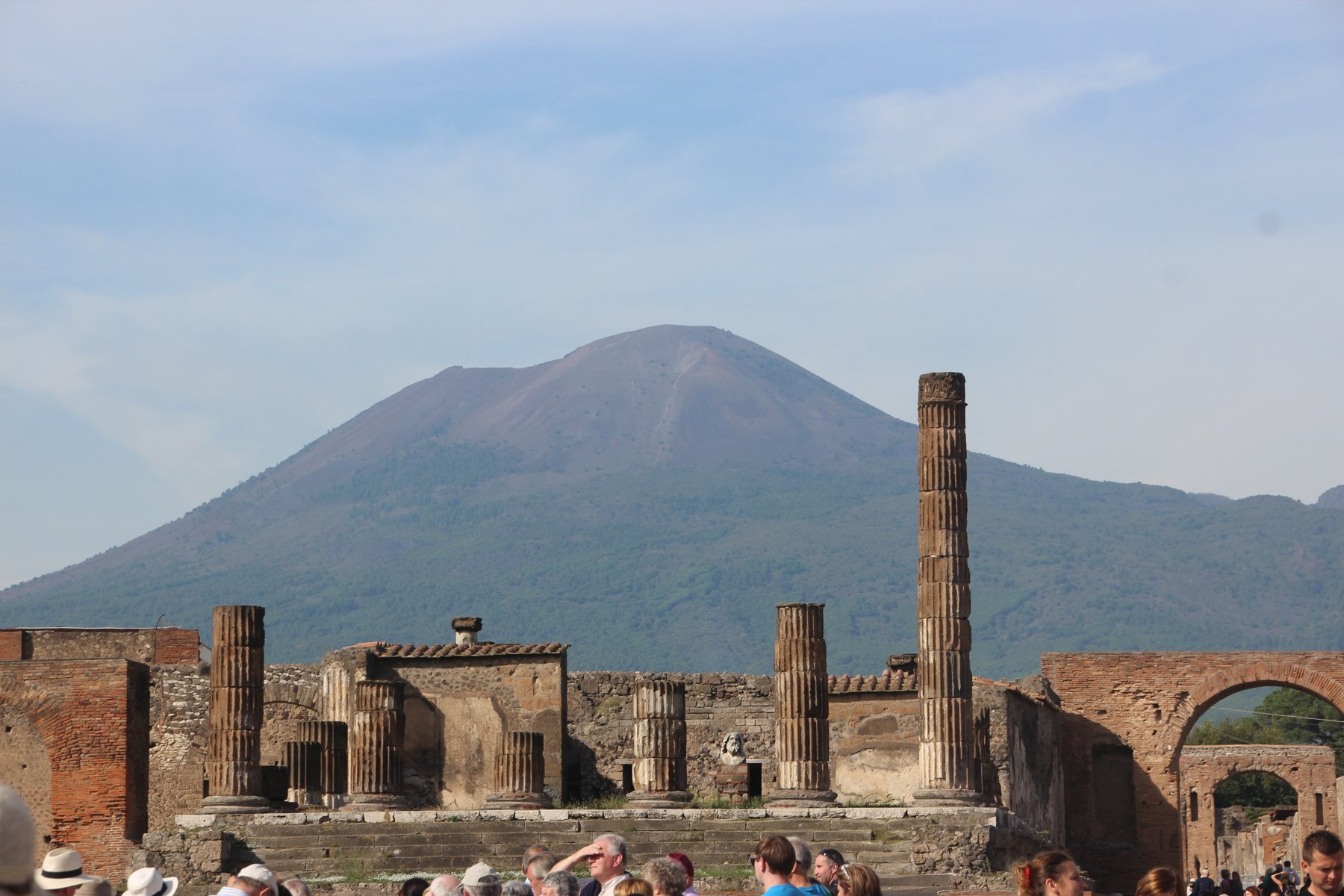 Columns and brick remains in Pompeii’s forum stand before the modern profile of Vesuvius, with visitors in the foreground.