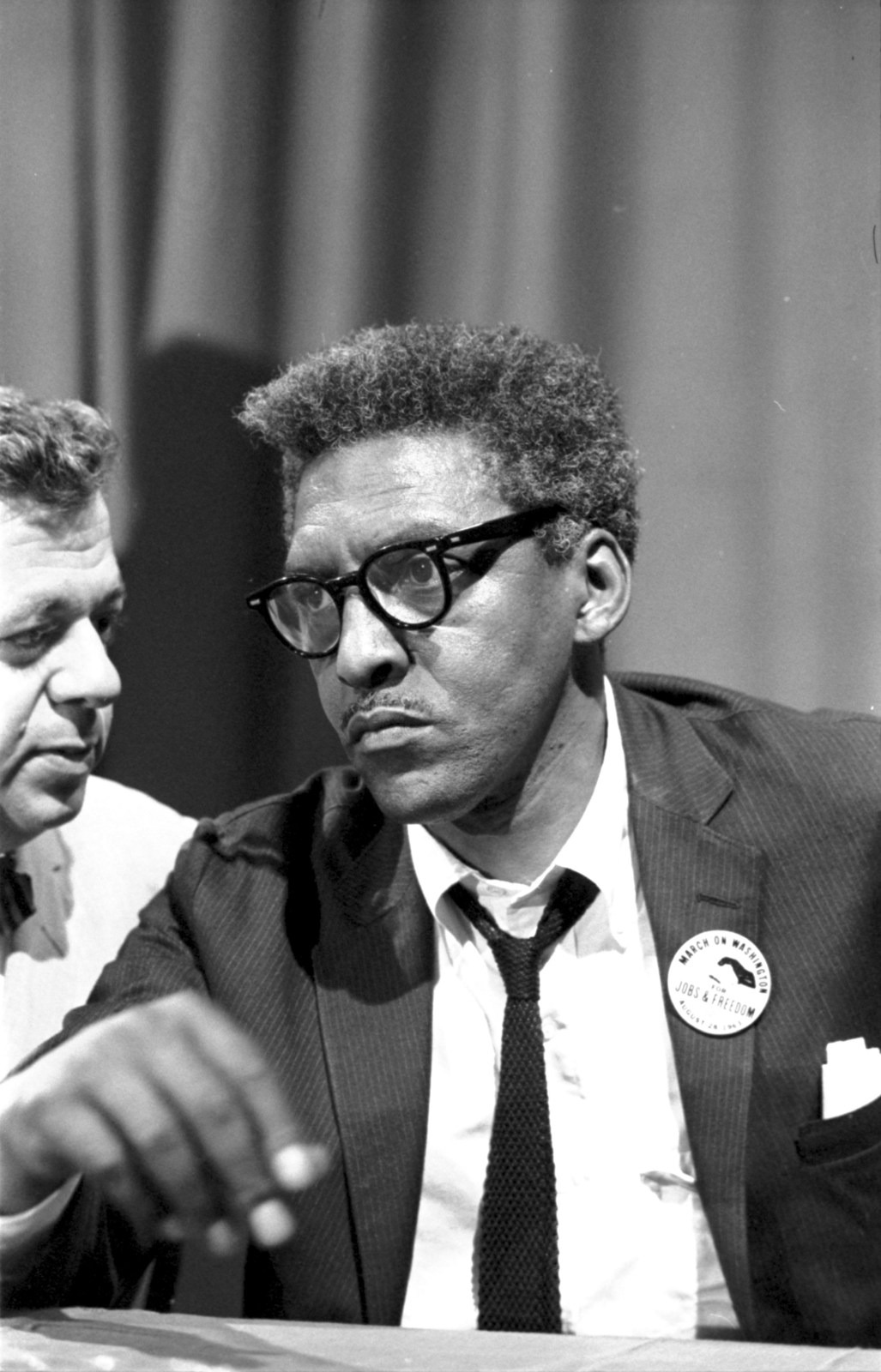 Bayard Rustin wears glasses and a March on Washington badge while seated at a news briefing.