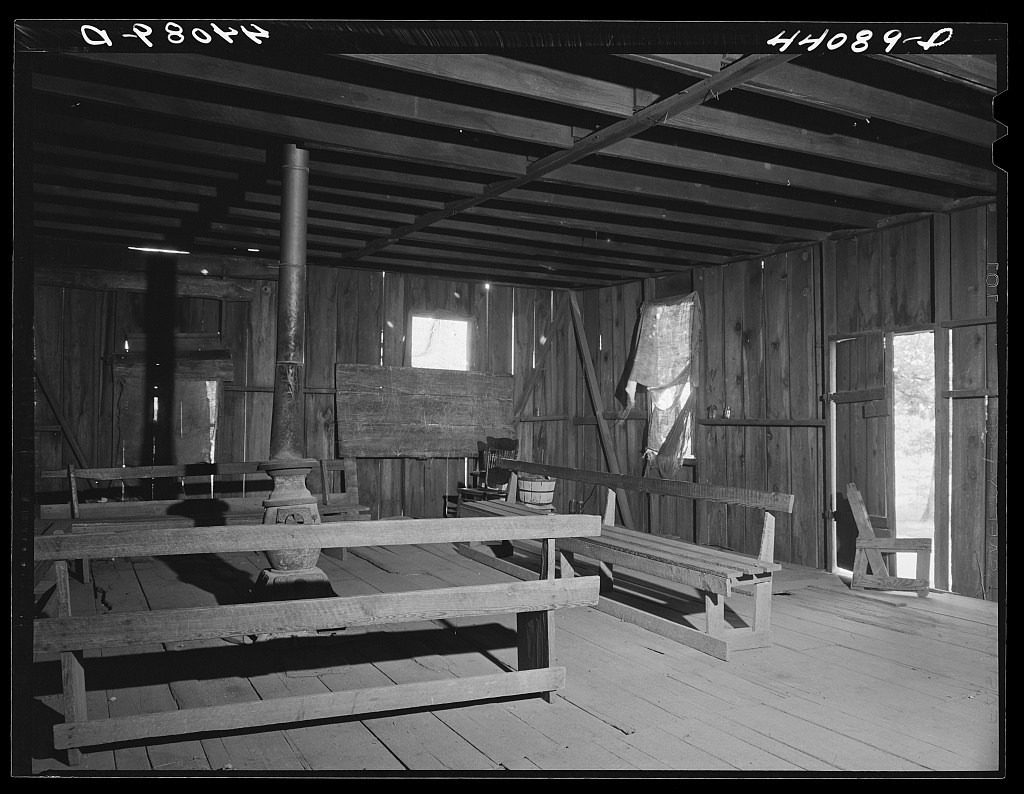 An empty wooden schoolroom contains benches, a stove, windows and a writing board.