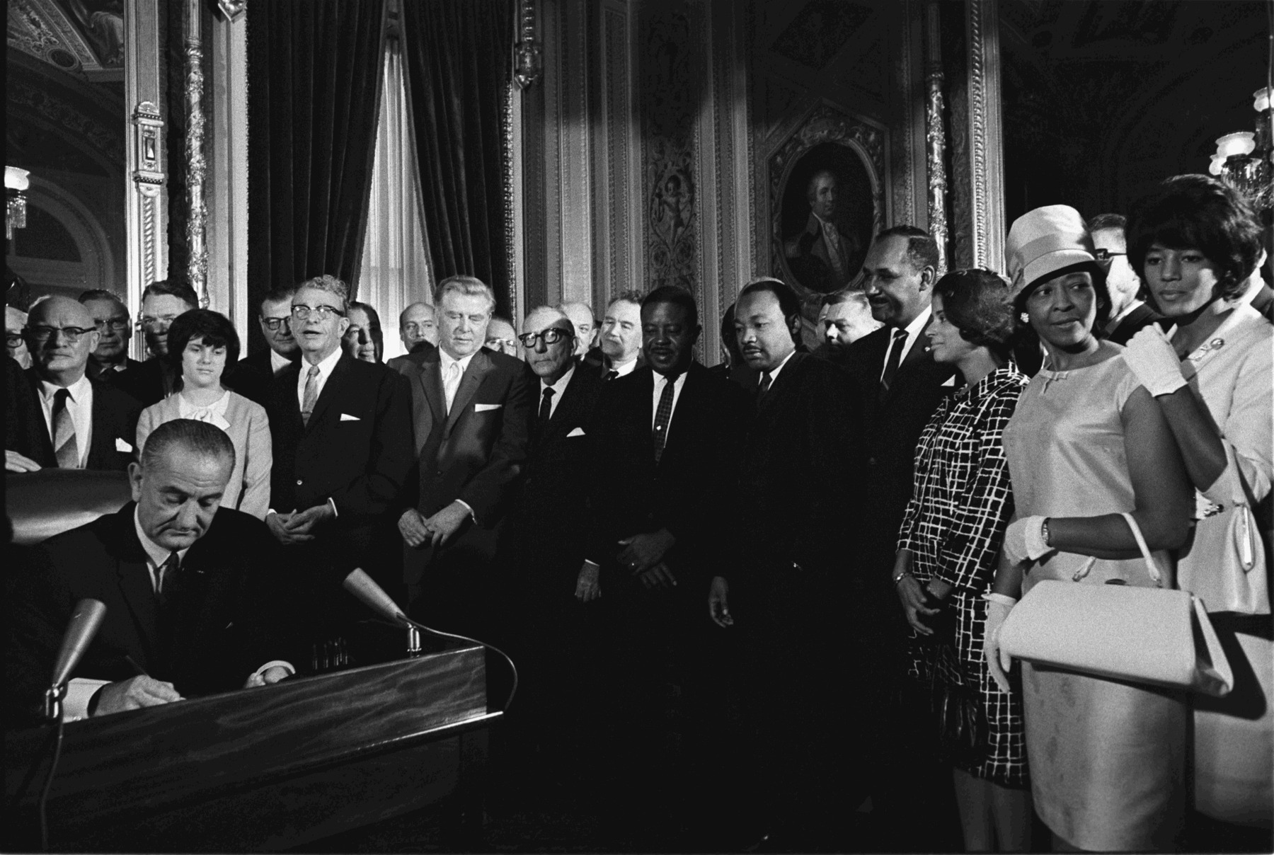Johnson sits at a desk signing a document while King and other attendees stand behind him.