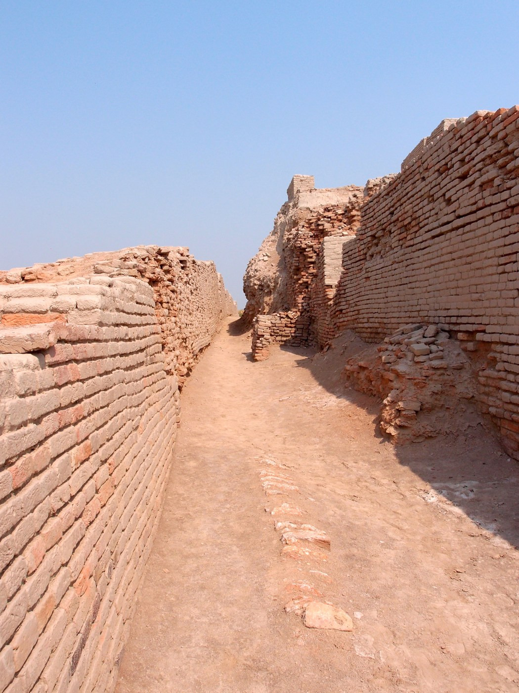 A narrow passage between exposed brick walls at Mohenjo-daro, with a visitor in the distance giving a sense of scale.