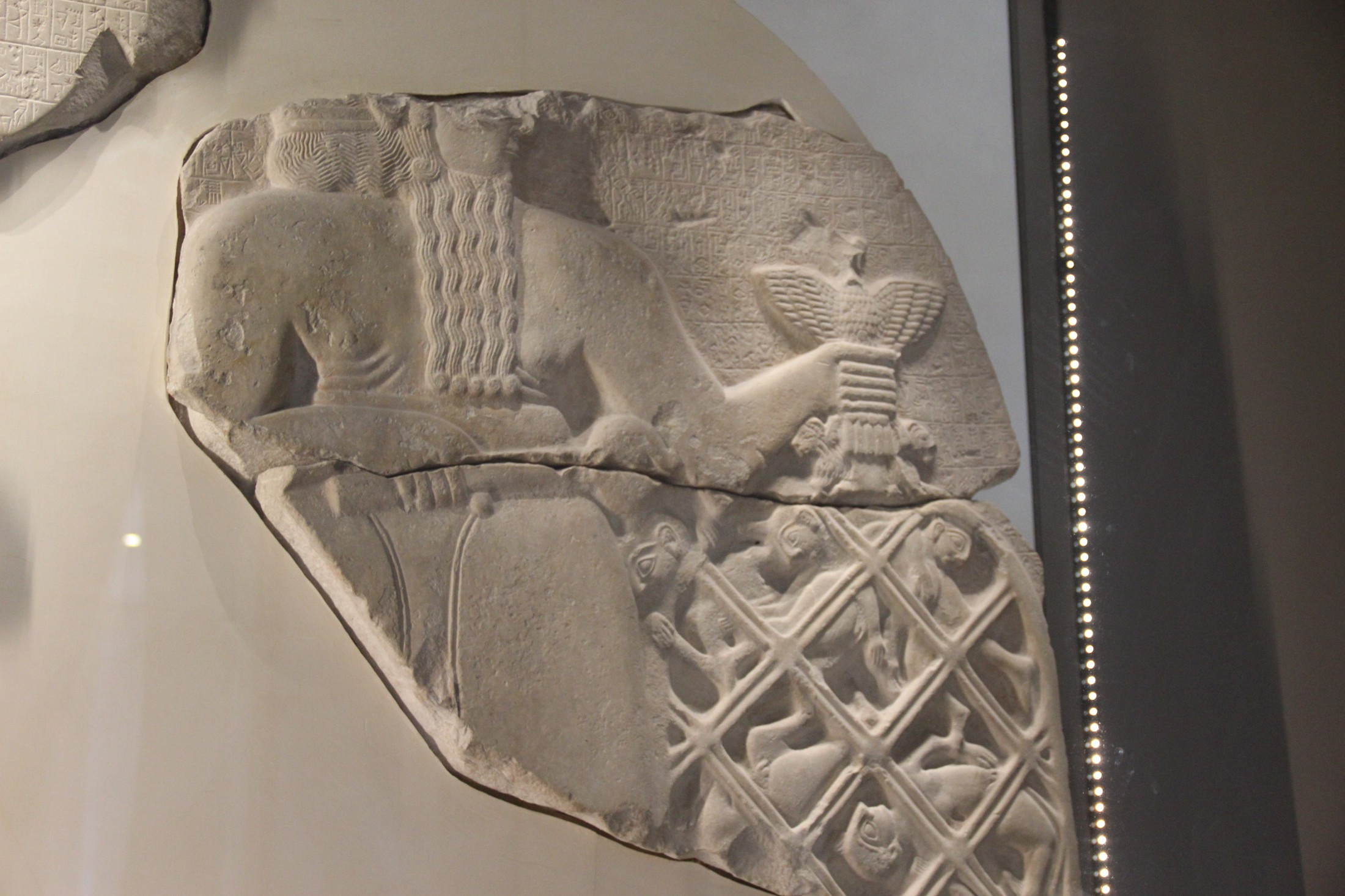 Surviving carved fragments show helmeted soldiers with spears and shields; pale modern supports indicate missing parts of the stele.